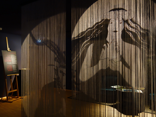 Das Bild zeigt eine schwach beleuchtete Kunstinstallation, bei der hängende Schnüre den Schatten eines großen Gesichts an die Wand werfen. Links ist ein Schild zu sehen.