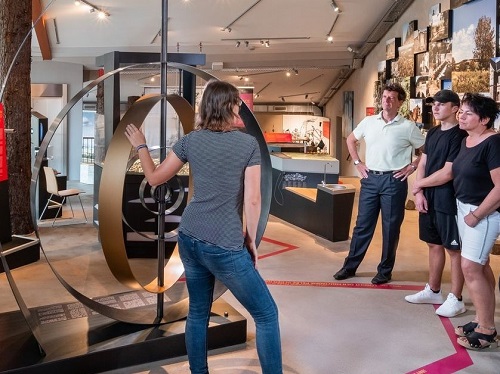 Eine Frau erklärt drei Personen in einem modernen Museum etwas und deutet dabei auf eine große, runde Metallskulptur.