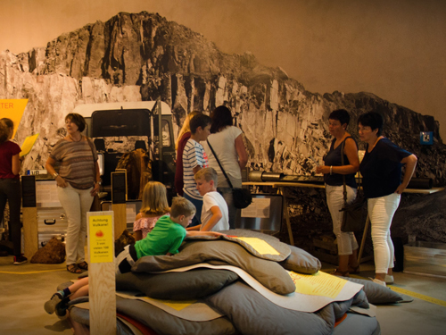 Mehrere Personen stehen in einem Ausstellungsraum vor einer großen Felswandkulisse, Kinder sitzen auf gepolsterten Sitzkissen auf dem Boden.