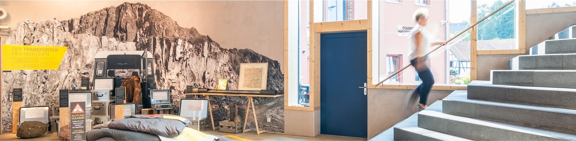 Ein modernes Interieur mit einem Wandbild, das eine Berglandschaft zeigt, sowie ausgestellter Kletterausrüstung und gerahmten Bildern. Eine Person steigt eine nahegelegene Treppe hinauf.