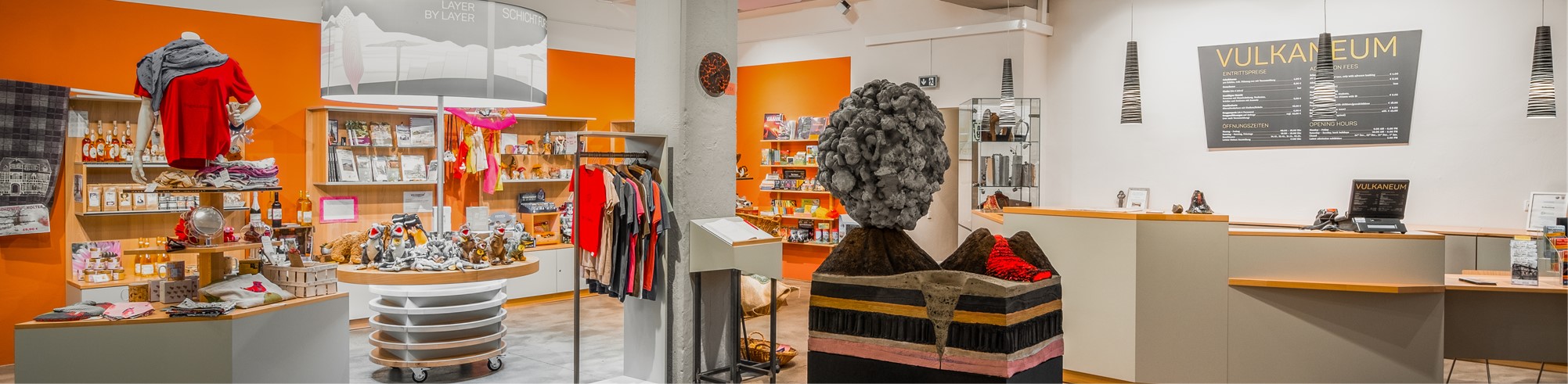 Ein lebhafter Museumsshop mit leuchtend orangefarbenen Wänden, in dem farbenfrohe Kleidung, Bücher und Souvenirs angeboten werden. Eine große, strukturierte Skulptur nimmt einen prominenten Platz ein.